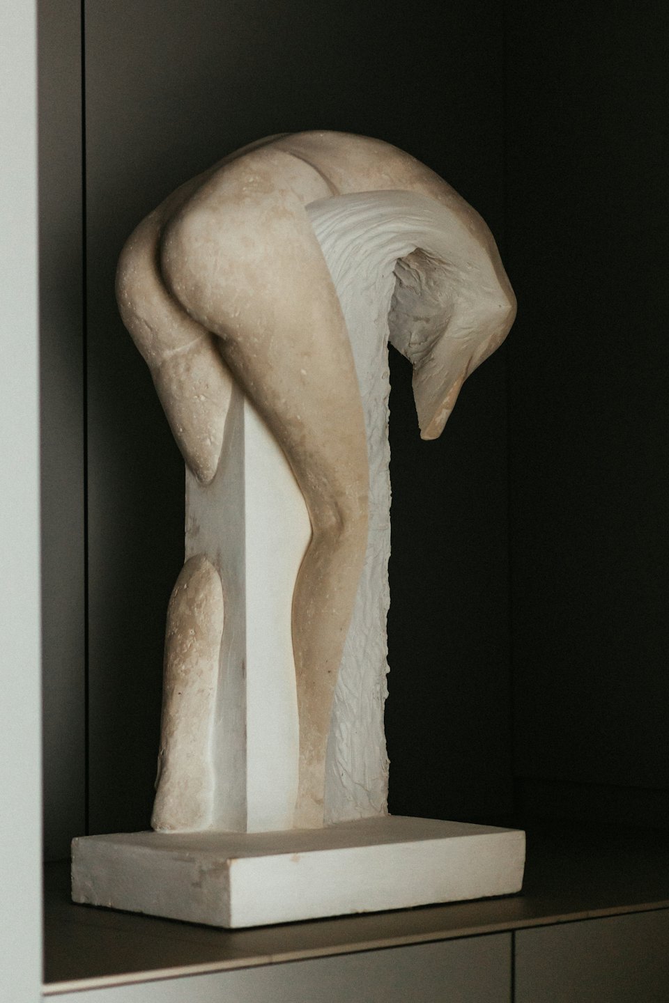 Strukturierte Steinskulptur einer gebeugten Figur auf einem weißen Sockel.